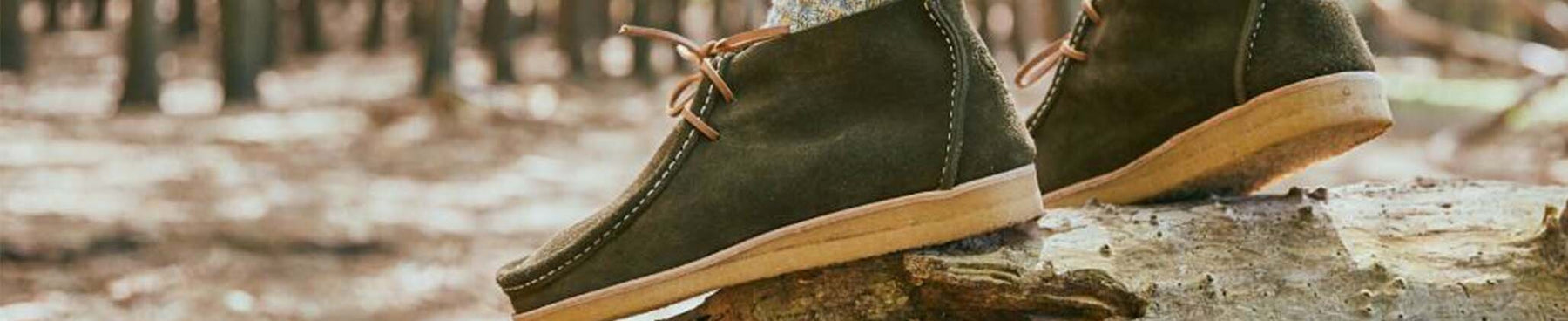 Clarks Originals Desert Boots in olive green suede with crepe soles, resting on a log in a forest, showcasing their rugged style and comfort for outdoor wear