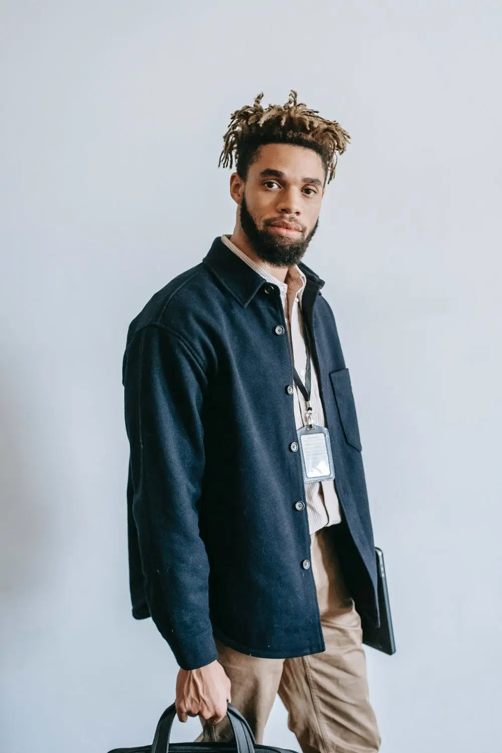 Stylish man with dreadlocks wearing a navy jacket overshirt, holding a black briefcase, against a light background, showcasing professional yet casual menswear.