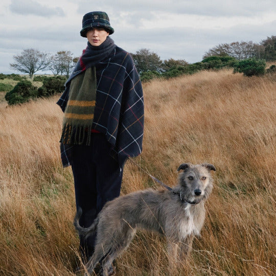 Person walking a dog in a field wearing a plaid Barbour coat, scarf, and hat. featured image 2