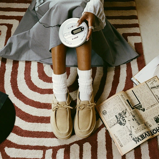 Person sitting on a patterned rug holding a white radio, wearing a gray skirt and Clarks Originals beige shoes. featured image 2