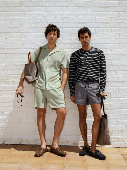 Two men in Paul Smith summer outfits with Signature Stripe tote bags, loafers, and relaxed tailoring against a white wall.