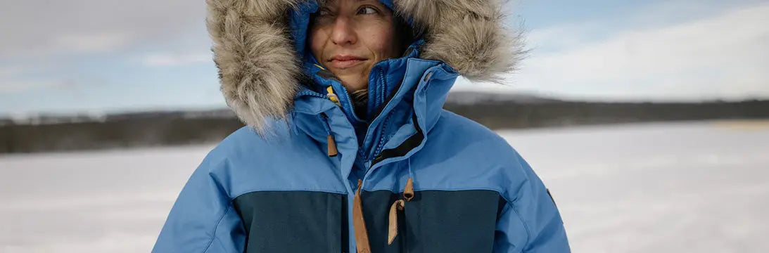 Person wearing a blue and turquoise Fjallraven winter jacket with a fur-lined hood, standing in a snowy landscape, evoking warmth and outdoor protection