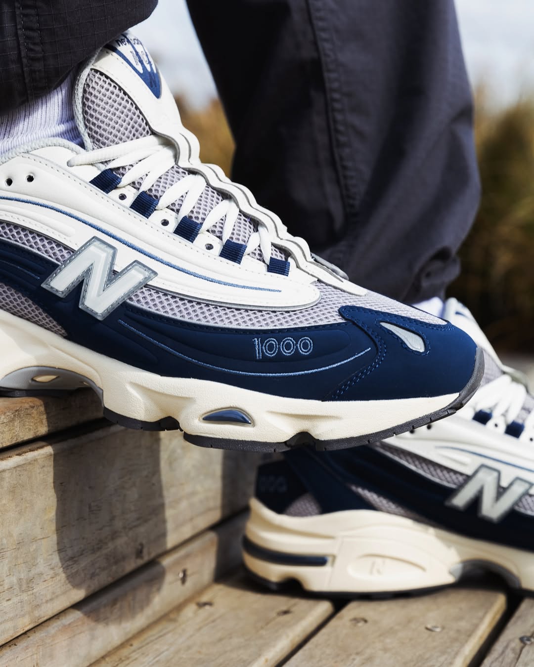 New Balance M1000 trainers in Navy and Sea Salt with mesh upper and reflective ‘N’ logo, worn outdoors on wooden steps.
