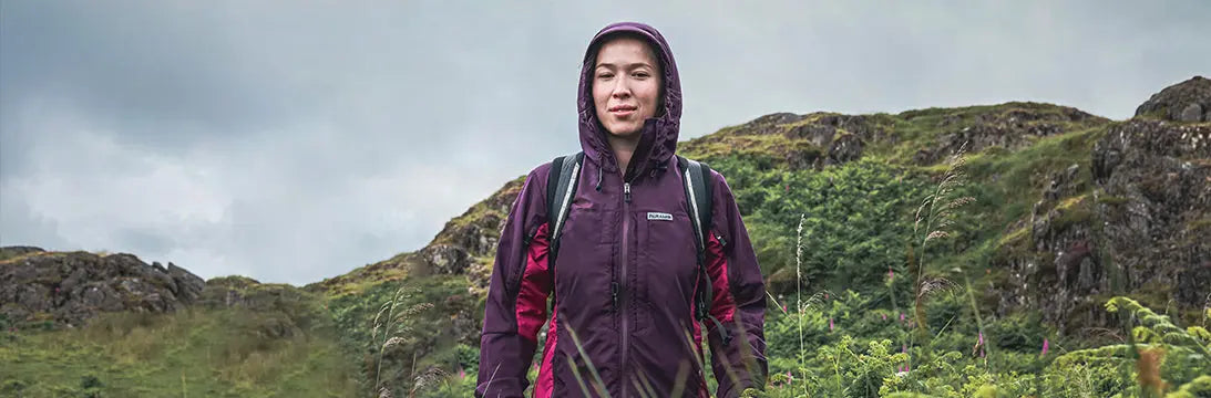 Hiker wearing a purple waterproof Paramo jacket with hood and backpack in a rugged mountain landscape, suggesting durable outdoor attire for hiking