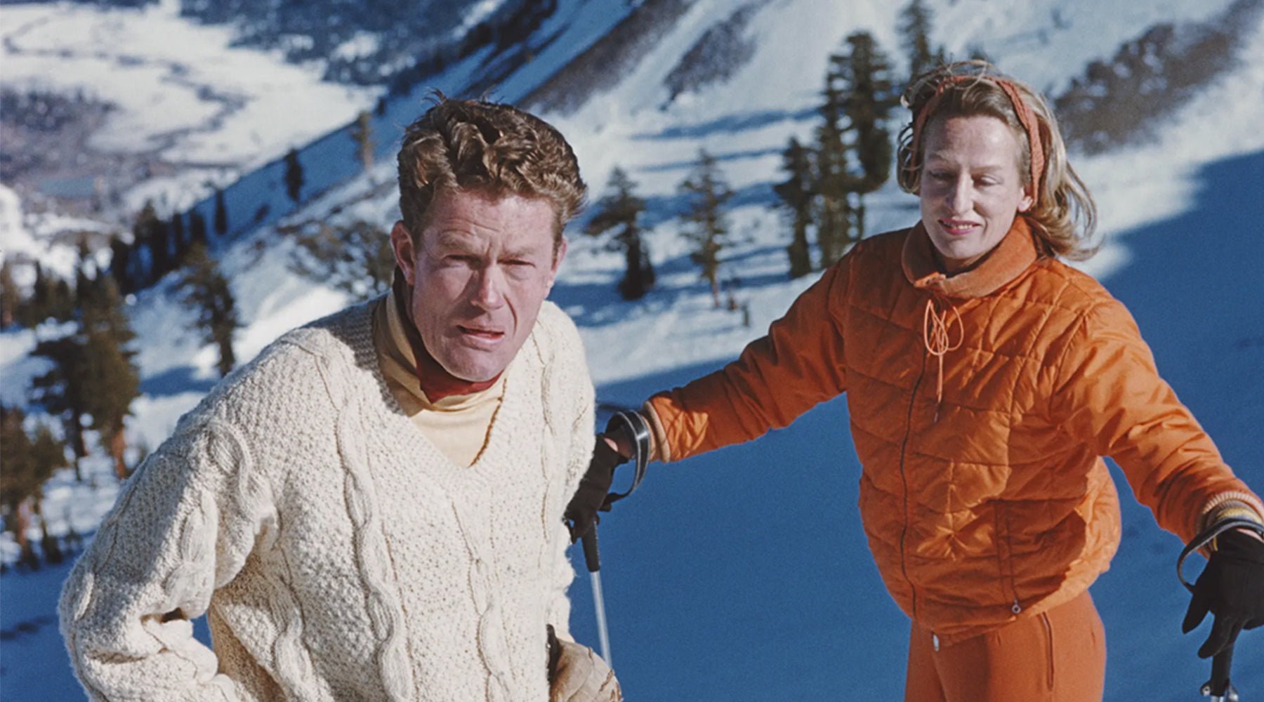 Two cross country skiers climbing a snowy mountain ridge wearing vintage knit jumper and orange retro ski jacket