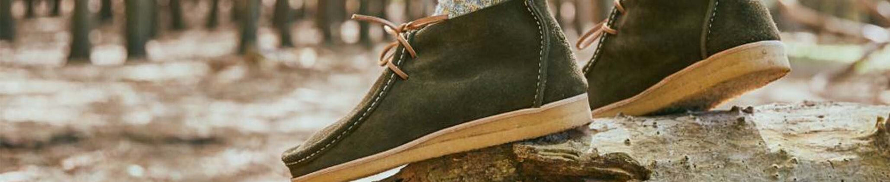 Clarks Originals Desert Boots in olive green suede with crepe soles, resting on a log in a forest, showcasing their rugged style and comfort for outdoor wear