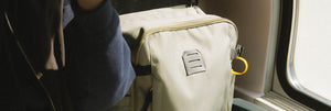 Cream-coloured commuter backpack with a modern design and distinctive branding, positioned next to a train window, highlighting on-the-go lifestyle accessories.