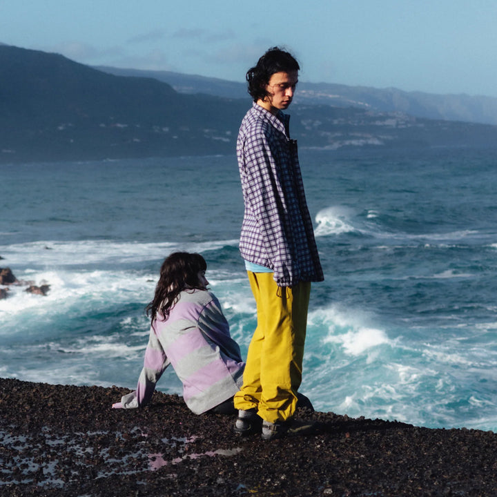 Two people standing on a rocky shore wearing colourfil outdoor clothing with ocean waves and mountains in the background