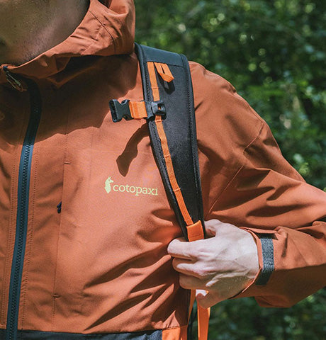 Close-up of a hiker adjusting the straps of a burnt orange Cotopaxi jacket, highlighting the brand's logo and the gear's functional design for outdoor activities.