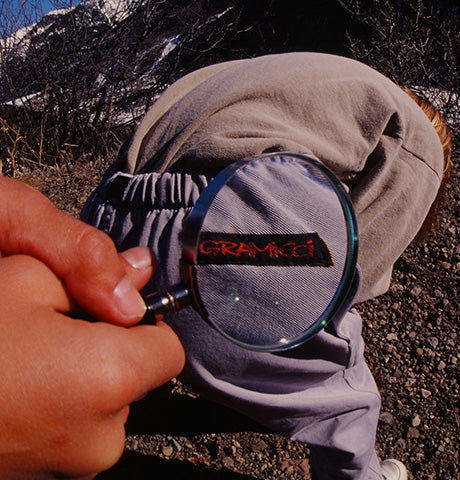 Hand holding a magnifying glass over a Gramicci label on a pair of grey outdoor trousers, highlighting the brand's detail and quality in adventure clothing.