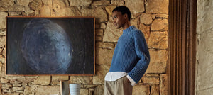 Woman in a chic blue ribbed Sunspel sweater and white shirt standing in a rustic room with a large abstract painting, exemplifying casual luxury womenswear