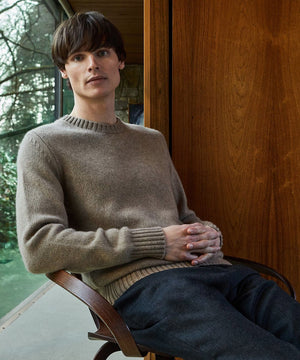 Man seated in a wooden chair wearing a classic oatmeal Sunspel crewneck sweater and dark trousers, set against a backdrop of large windows and natural scenery.