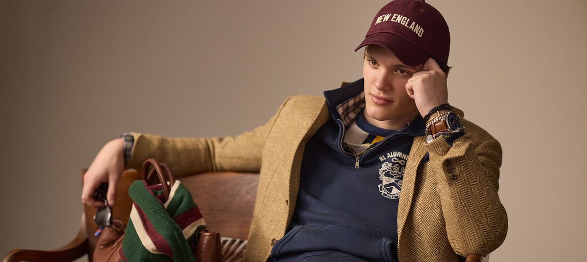 Man wearing a tan Polo Ralph Lauren jacket and maroon cap with 'New England' text, sitting on a chair.